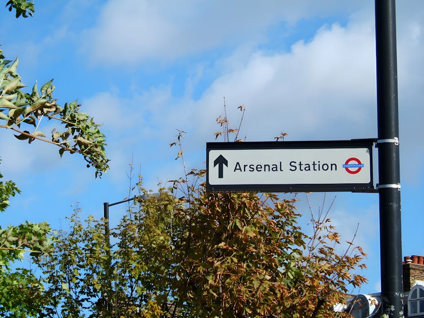 A rectangular road sign attached to a black metal pole indicating the direction to 'Arsenal Station' with a black upward arrow. The sign has a white background, black text, and features the red, white, and blue London Underground roundel logo on the right side. Behind the sign, there are trees with green and reddish-brown leaves, some branches extending into the frame. The sky above is bright blue with scattered white clouds, and parts of rooftops with chimney pots are visible at the bottom right. The overall scene is outdoors in a residential or urban area. This image could be relevant to discussions on local infrastructure, transport options, or independent ways of handling waste and rubbish collection near popular landmarks or public transport hubs in areas like Islington.