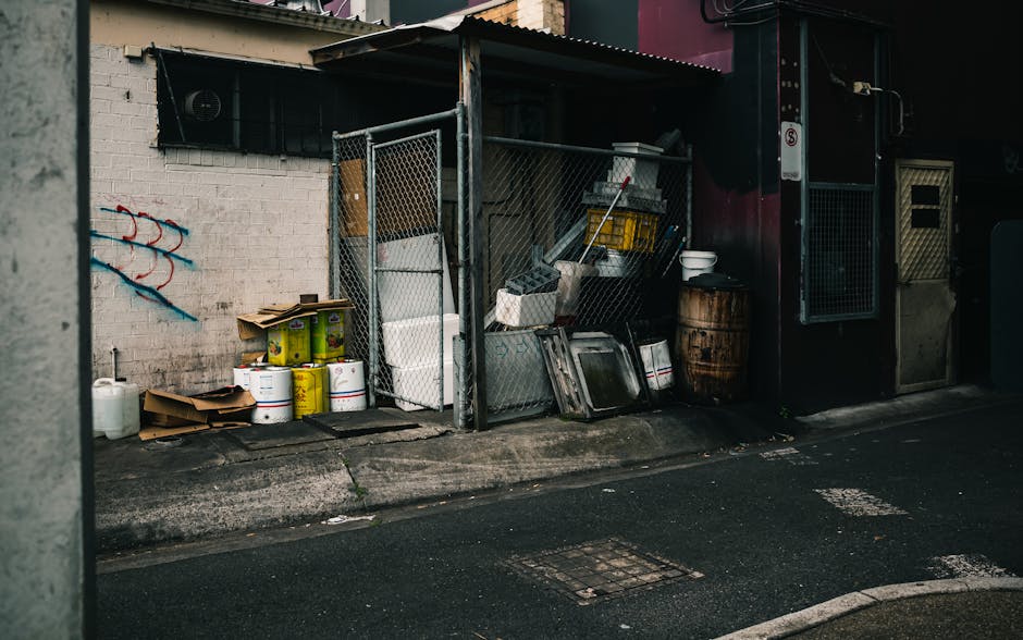 A small outdoor storage area situated beside a building is filled with various discarded items and waste materials. The enclosure is constructed with a metal frame and chain-link fencing, providing a partial barrier around the stored objects. Inside, there are several white plastic buckets, some with yellow labels, stacked against a white brick wall, which features a small window with a black ventilation cover. Adjacent to the buckets, there are flattened cardboard boxes, and nearby a large white plastic container. Leaning against the fencing are metal trays, a yellow container, and other miscellaneous debris. To the right, there is an aged wooden barrel, and on a shelf inside the enclosure, various small items and tools are visible. The scene is illuminated by natural daylight, capturing the gritty urban environment on a paved street with a drainage grate in the foreground. This image reflects a typical private waste storage area, often associated with independent rubbish collection and clearance services provided by companies like House Clearance Islington for efficient rubbish removal and alternative waste handling in urban settings.