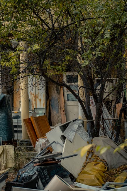 In the foreground, a disorganized pile of rubbish is scattered, including white and beige appliances with a textured finish, such as a washing machine and possibly an oven, partially covered by grey and yellow plastic bags. Some smaller objects like black plastic containers and metal tools are also visible among the debris. Behind the pile, a lush, leafy tree with green foliage extends its branches over the rubbish, partially obscuring an aged building with a weathered facade, featuring rusted metal panels and peeling paint. To the left, a dark tarpaulin or outdoor cover is draped over an object or structure, while on the right, a metal fence is partially visible. The scene is outdoors, set on a paved or concrete surface with natural, diffuse daylight illuminating the scene, emphasizing the contrast between the clutter and the surrounding greenery. This image from House Clearance Islington highlights a cluttered area awaiting clearance, typical of scenes where independent collection or private waste handling is involved, emphasizing the need for professional rubbish removal services near Angel Station N1.