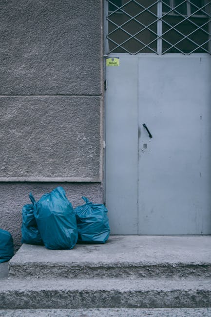 In the foreground, a disorganized pile of rubbish is scattered, including white and beige appliances with a textured finish, such as a washing machine and possibly an oven, partially covered by grey and yellow plastic bags. Some smaller objects like black plastic containers and metal tools are also visible among the debris. Behind the pile, a lush, leafy tree with green foliage extends its branches over the rubbish, partially obscuring an aged building with a weathered facade, featuring rusted metal panels and peeling paint. To the left, a dark tarpaulin or outdoor cover is draped over an object or structure, while on the right, a metal fence is partially visible. The scene is outdoors, set on a paved or concrete surface with natural, diffuse daylight illuminating the scene, emphasizing the contrast between the clutter and the surrounding greenery. This image from House Clearance Islington highlights a cluttered area awaiting clearance, typical of scenes where independent collection or private waste handling is involved, emphasizing the need for professional rubbish removal services near Angel Station N1.