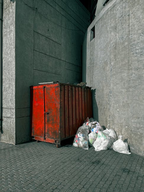 A weathered red metal skip with visible rust and peeling paint is positioned at the corner of a building’s exterior, adjacent to a grey concrete wall and dark green panels. The skip is partially overflowing, with several transparent plastic bags filled with mixed rubbish, including plastic bottles and paper, leaning against its side on the ground. The surface beneath the skip is made of dark interlocking paving stones, which extend across the paved area. A narrow vertical window is visible high on the grey wall behind the skip, and the scene is lit by natural daylight, casting soft shadows. The setting appears to be an urban alley or service yard, reflecting a typical environment where private rubbish collection or on-site clearance might take place, separate from council waste disposal services. This image emphasizes the types of waste handled by independent waste management services such as those offered by House Clearance Islington.