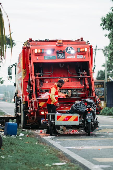 A worker dressed in a high-visibility orange and yellow vest is operating a large red rubbish collection vehicle, with its rear compactor open and some debris visible inside. The vehicle is parked on the side of a paved street, with a small black motorcycle secured nearby on a stand, partially blocking the vehicle's rear area. The scene includes a black plastic bag placed on top of the motorcycle, visible nearby as part of waste collection or disposal. In the background, there are trees, utility poles with wires, and a building with signage, indicating an urban or suburban environment. The sky appears overcast, providing diffuse natural lighting that highlights the textures of the vehicle's metallic surface, the rough, weathered asphalt, and the surrounding greenery. The scene captures a moment of private or independent waste handling, often associated with rubbish removal services such as those provided by House Clearance Islington, which may support alternative waste collection methods outside local authority rubbish collection schemes.