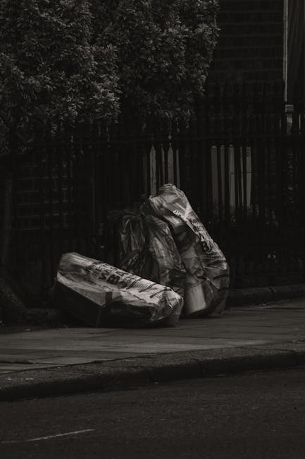 A black and white photograph showing three large plastic rubbish bags placed on the pavement beside a dark wooden fence and a brick wall, adjacent to a sidewalk. The bags appear to contain waste materials and are slightly crumpled with visible creases and compression marks. The background includes dense foliage of trees or bushes and part of a building behind the fence, suggesting an urban or suburban setting. The scene depicts an external waste collection point, likely related to a private rubbish disposal service such as House Clearance Islington, aligning with the context of alternative rubbish collection and on-site waste handling. The natural lighting creates subtle shadows across the bags and pavement, emphasizing their texture and volume, suitable for an article on rubbish removal options in the local area.