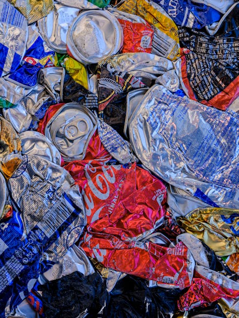 Close-up view of a pile of crushed aluminum cans and aluminum foil wrappers, mainly in silver, red, blue, and gold colors, with some identifiable brand labels such as Coca-Cola visible amidst the crumpled metallic debris. The cans are tightly compressed and layered, with sharp edges and textured surfaces reflecting light, indicating recent disposal. Surrounding the crushed cans, there are additional aluminum packaging and fragments, creating a chaotic assembly of waste materials typical of collection from household or commercial rubbish collection for recycling or private waste removal. The scene appears to be set outdoors or in a designated waste area, with no background elements visible, focusing entirely on the metallic rubbish. The overall composition emphasizes the type of recyclable waste material often handled by independent rubbish removal services like House Clearance Islington, which specialise in alternative waste handling and on-site clearance of such household or commercial waste items.