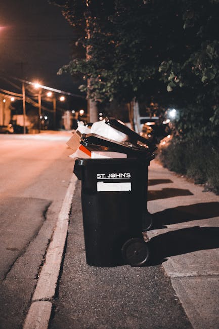 A black wheelie bin with the label 'St. John's' positioned on a street sidewalk at night, filled with a variety of rubbish including cardboard boxes, plastic containers, and paper waste. The bin is situated near the curb, with the waste visibly overflowing, some items leaning out and partially resting atop the lid. The scene is illuminated by streetlights casting a warm glow, with shadows stretching across the pavement and nearby shrubbery. In the background, there are streetlights, a tree, and parked vehicles, indicating an urban residential area. The environment suggests an instance of private waste disposal reflecting alternative rubbish collection methods, possibly handled by a local waste management service such as House Clearance Islington, or similar providers, emphasizing the importance of proper rubbish removal and disposal.