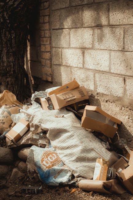 A small pile of mixed waste materials is situated against a rough, beige brick wall outdoors. The collection includes several flattened cardboard boxes, some partially open, revealing their plain brown interiors, with printed labels and barcodes visible on their surfaces. There are also large, tightly wrapped white plastic or fabric bags, possibly filled with more rubbish, placed on top of or near the cardboard. The waste appears to be discarded household or retail packaging, with textures varying from smooth cardboard to crinkled paper and soft fabric-like coverings. The pile rests on a ground surface comprised of small dark stones or gravel, with a portion of a nearby tree trunk visible on the left side of the image. The scene is lit naturally, suggesting daytime, with no signs of active collection or loading. This cluttered accumulation demonstrates a typical scenario where an independent waste collection or private disposal service, such as House Clearance Islington, might be called to manage such rubbish removal, especially when local authority collections are unavailable or unsuitable for bulky waste.