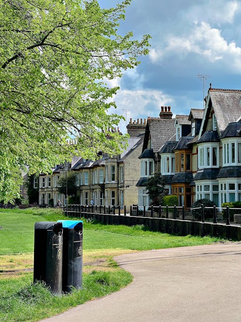 A row of traditional Victorian-style terraced houses with varied front façades, including bay windows and dormer windows on the rooftops, situated along a residential street. In the foreground, two black public refuse bins are positioned on a grassy verge beside a paved footpath, with some scratches and wear visible on their surfaces. A leafy tree with bright green foliage extends over the scene on the left side, partially shading the bins and the grass beneath. Behind the bins, a short wooden fence runs parallel to the houses, enclosing small front gardens with neatly maintained shrubs and plants. Above, the sky is partly cloudy with patches of blue visible through white and gray clouds, creating natural daylight conditions that highlight the textures of the brickwork, painted woodwork, and roof tiles. The environment suggests a peaceful, well-established residential area, where private waste disposal and potential arrangements for independent rubbish collection through local waste management services like House Clearance Islington may be relevant for maintaining cleanliness and disposal of household waste.