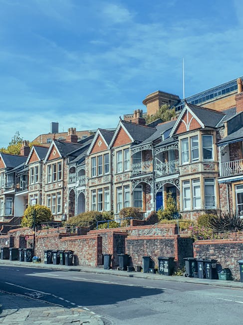 A row of traditional Victorian-style terraced houses with varied front façades, including bay windows and dormer windows on the rooftops, situated along a residential street. In the foreground, two black public refuse bins are positioned on a grassy verge beside a paved footpath, with some scratches and wear visible on their surfaces. A leafy tree with bright green foliage extends over the scene on the left side, partially shading the bins and the grass beneath. Behind the bins, a short wooden fence runs parallel to the houses, enclosing small front gardens with neatly maintained shrubs and plants. Above, the sky is partly cloudy with patches of blue visible through white and gray clouds, creating natural daylight conditions that highlight the textures of the brickwork, painted woodwork, and roof tiles. The environment suggests a peaceful, well-established residential area, where private waste disposal and potential arrangements for independent rubbish collection through local waste management services like House Clearance Islington may be relevant for maintaining cleanliness and disposal of household waste.