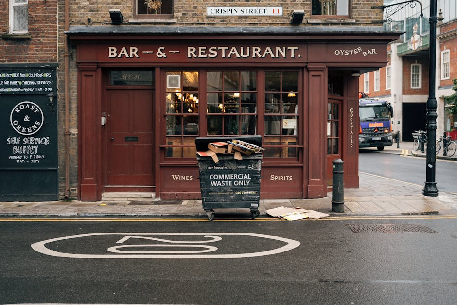 In front of a brown-painted building housing a bar and restaurant with large glass windows, a black commercial waste bin is positioned on the sidewalk, filled with cardboard boxes and paper waste, some spilling onto the pavement nearby. The bin is labeled 'COMMERCIAL WASTE ONLY' in white lettering. To the left, there is a blackboard sign advertising a self-service buffet, and to the right, a black bollard protects the corner of the building. Behind the bin, a white-painted road marking indicates a no-parking zone. A black street lamp and several parked bicycles are visible further along the street, with a black truck parked on the road beyond the utility pole. The building features traditional brickwork topped with a sign for 'CRISPIN STREET' above the storefront. The scene suggests a typical urban setting where private waste collection services might be employed for commercial rubbish disposal, echoing themes of alternative waste handling outside of local authority collection methods, in line with professional rubbish removal practices of House Clearance Islington.
