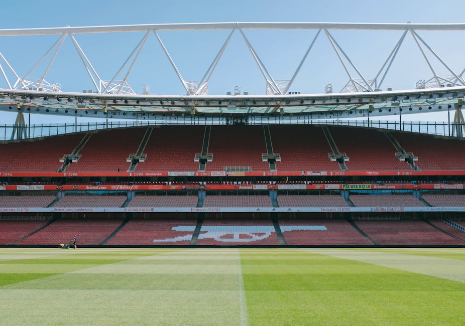 A wide view of the interior of a large, modern football stadium with a well-maintained grass pitch in the foreground, displaying varied shades of green in the freshly mowed turf. The seating areas, predominantly red, stretch across multiple tiers and are mostly unoccupied, with a few small figures visible in the distance. The stadium's infrastructure includes a prominent, white steel roof structure supported by trusses and cables, arcing high above the seating sections, with the sky visible in the background. Along the sides of the seating tiers, digital advertising boards run continuously, some displaying text and logos. The overall lighting is natural daylight, casting soft shadows and highlighting the vibrant contrast between the green playing surface and the red seating areas. While no rubbish or waste materials are visible, the image's clean environment exemplifies well-maintained facilities aligned with professional sports venues, unrelated to rubbish removal services.