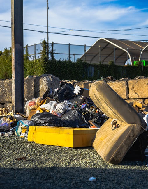 In front of a brown-painted building housing a bar and restaurant with large glass windows, a black commercial waste bin is positioned on the sidewalk, filled with cardboard boxes and paper waste, some spilling onto the pavement nearby. The bin is labeled 'COMMERCIAL WASTE ONLY' in white lettering. To the left, there is a blackboard sign advertising a self-service buffet, and to the right, a black bollard protects the corner of the building. Behind the bin, a white-painted road marking indicates a no-parking zone. A black street lamp and several parked bicycles are visible further along the street, with a black truck parked on the road beyond the utility pole. The building features traditional brickwork topped with a sign for 'CRISPIN STREET' above the storefront. The scene suggests a typical urban setting where private waste collection services might be employed for commercial rubbish disposal, echoing themes of alternative waste handling outside of local authority collection methods, in line with professional rubbish removal practices of House Clearance Islington.